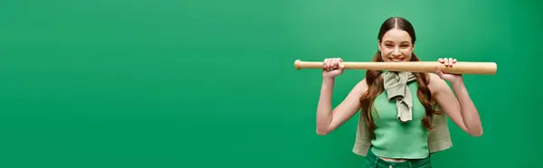 Uma jovem, bela mulher de 20 anos tem um taco de beisebol na frente de seu rosto em um estúdio em verde. — Fotografia de Stock