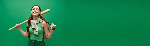 A young beautiful woman in her 20s confidently holding a baseball bat against a vibrant green background. — Stock Photo