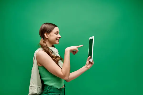 Una giovane donna sulla ventina tiene un tablet e lo indica in un ambiente da studio con uno sfondo verde. — Foto stock