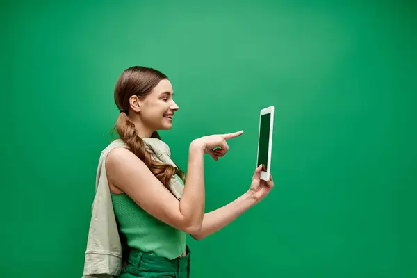 A young woman in her 20s holds a tablet and points at it in a studio setting with a green background. — Stock Photo