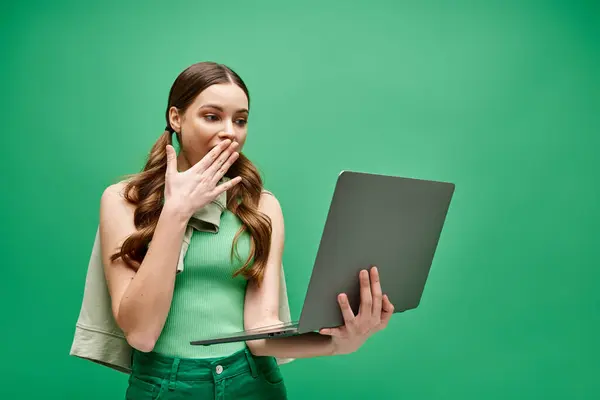 A young woman in her 20s covers her mouth while using a laptop in a studio setting, hinting at hidden thoughts or emotions. — Stock Photo