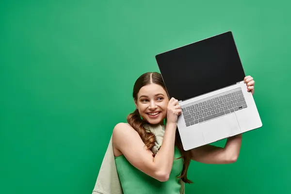 A woman in her 20s holds a laptop up to her face in a studio setting against a green background. — Stock Photo