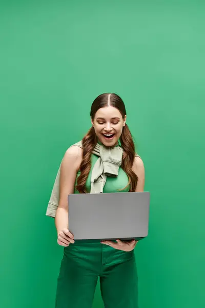 A young woman in her 20s stands in front of a green wall, confidently holding a laptop. — Stock Photo