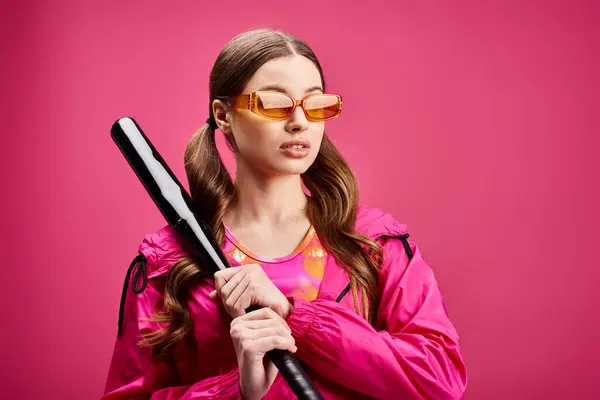 A stylish woman in her 20s, wearing a pink jacket, confidently holds a baseball bat against a vibrant pink background. — Stock Photo