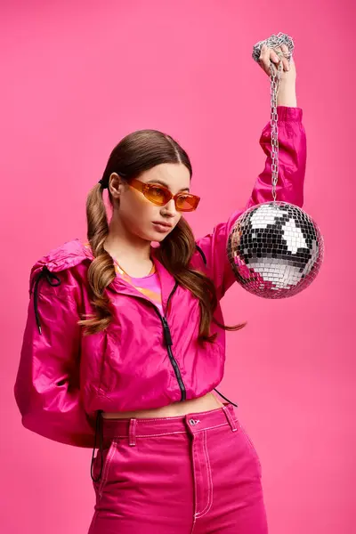 A stylish young woman in her 20s dressed in a vibrant pink outfit, holding a shimmering disco ball in a studio setting with a pink background. — Stock Photo