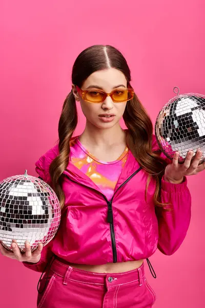 Stylish woman in her 20s, wearing a pink jacket, holds two shimmering disco balls in a vibrant studio setting. — Stock Photo