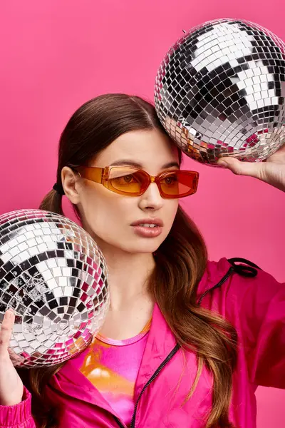 Uma jovem elegante com 20 anos, usando óculos de sol, segurando alegremente duas bolas de discoteca em um estúdio com um fundo rosa. — Fotografia de Stock