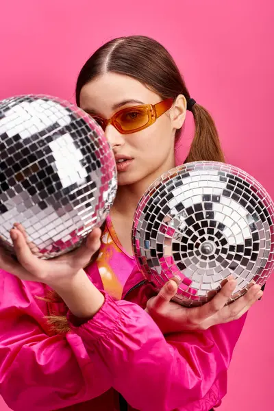 Una mujer joven y elegante de unos 20 años sostiene dos bolas de discoteca en un estudio con un fondo rosa, exudando un ambiente glamuroso. - foto de stock