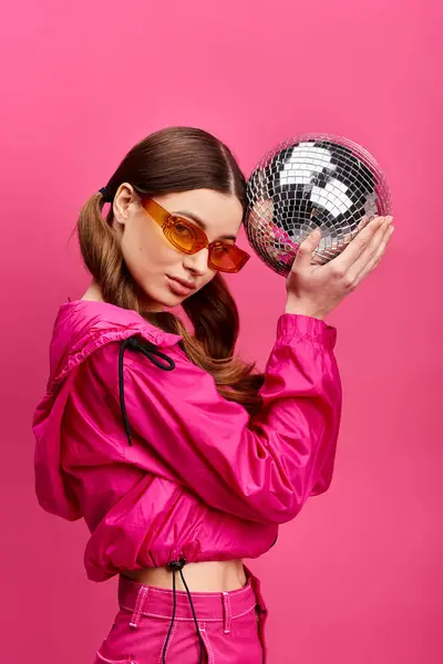 Una donna elegante sulla ventina che indossa una giacca rosa, che tiene una scintillante palla da discoteca in uno studio con uno sfondo rosa. — Foto stock