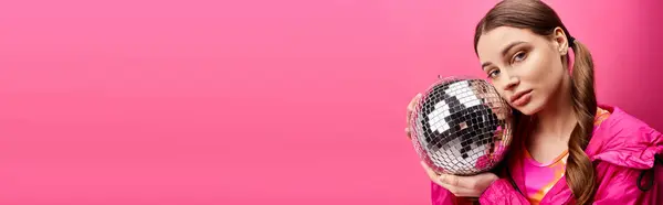 Junge Frau in den Zwanzigern hält Discokugel vor ihr Gesicht und schafft ein skurriles und geheimnisvolles Spektakel vor rosa Hintergrund. — Stockfoto