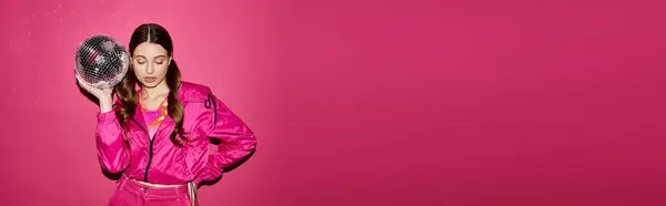 A stylish woman in her 20s, wearing a pink jacket, poses gracefully while holding a disco ball against a vibrant pink backdrop. — Stock Photo