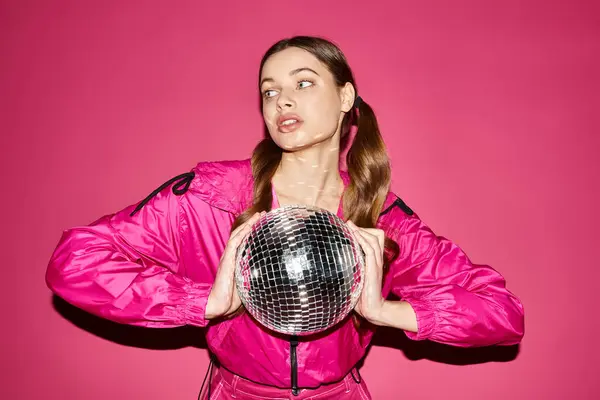 A stylish woman in her 20s wearing a pink jacket holding a glittering disco ball in front of a vibrant pink background. — Stock Photo