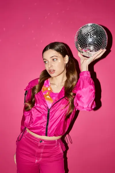 A stylish woman in her 20s, wearing a pink jacket, holding a disco ball in a studio with a pink background. — Stock Photo