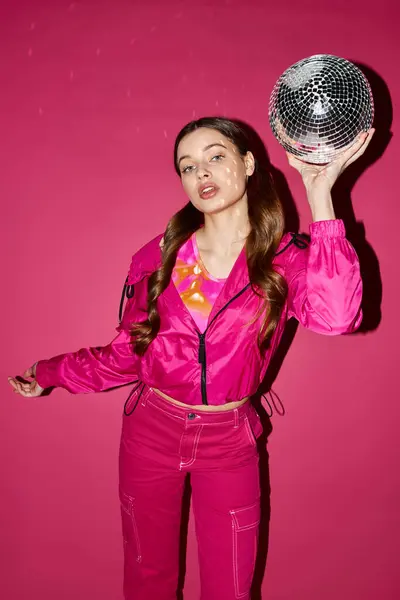 Una mujer de 20 años con estilo sostiene una bola de discoteca en su cara en un estudio con un fondo rosa, creando un reflejo deslumbrante. - foto de stock