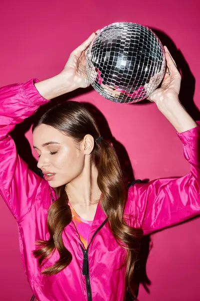 A stylish young woman in her 20s wearing a pink jacket, holding a disco ball in front of a pink background. — Stock Photo