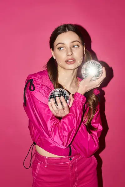 A stylish young woman in her 20s, dressed in a pink outfit, delicately holding a silver ball against a pink background. — Stock Photo