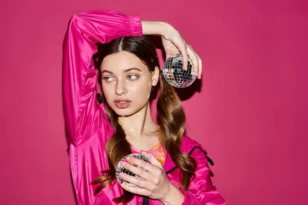 A stylish young woman in her 20s wearing a pink outfit holds a dazzling disco ball in a studio with a pink background. — Stock Photo