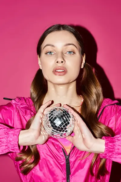 Une femme élégante dans la vingtaine tient une boule disco, rayonnant de lumières colorées, dans un studio vibrant avec un fond rose. — Photo de stock