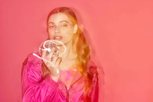 Una joven con un vestido rosa sostiene elegantemente una bola de espejo, creando un ambiente soñador y mágico en un estudio con un fondo rosa. - foto de stock
