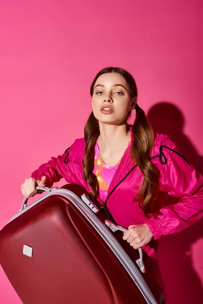 A stylish young woman in her 20s, wearing a pink jacket, holding a red suitcase against a pink backdrop. — Stock Photo