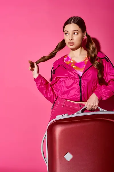 Une jeune femme élégante dans la vingtaine portant une veste rose tenant une valise rouge dans un décor de studio avec un fond rose. — Photo de stock
