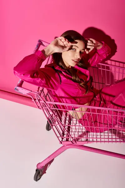 A stylish young woman in her 20s wearing a pink dress, sitting inside a pink shopping cart in a studio with a pink background. — Stock Photo