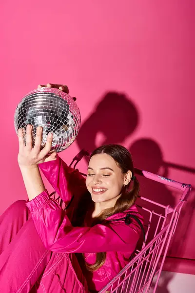 Uma jovem mulher em uma roupa rosa elegante segurando uma bola de discoteca, exalando glamour e diversão contra um fundo rosa vibrante. — Fotografia de Stock