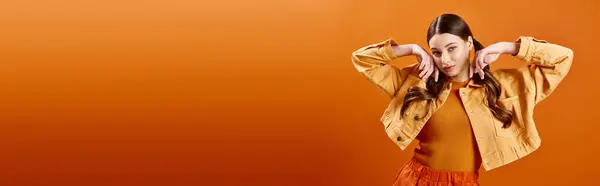 Young woman in her 20s wearing a bright yellow dress with her hands on her head in a studio setting against an orange background. — Stock Photo