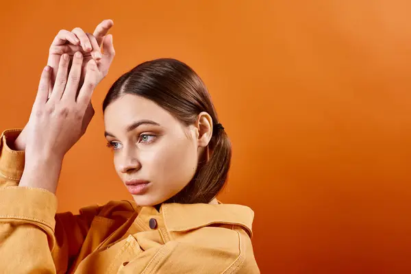 A stylish woman in her 20s raises her hands in a yellow jacket against an orange backdrop in a studio setting. — Stock Photo