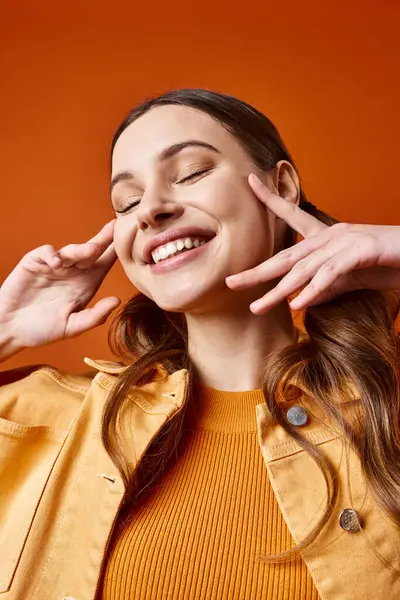 A stylish woman in her 20s smiling while delicately touching her ear with her hands against an orange studio backdrop. — Stock Photo