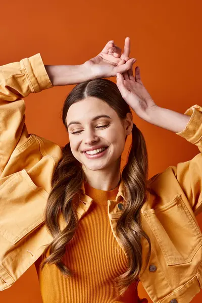 A young stylish woman in her 20s with a yellow shirt, joyfully raising her hands above her head against an orange background. — Stock Photo