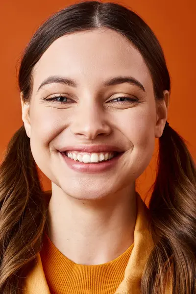 Uma jovem elegante na casa dos 20 anos com cabelos longos está sorrindo brilhantemente, vestindo uma camisola amarela vibrante contra um pano de fundo laranja.. — Fotografia de Stock