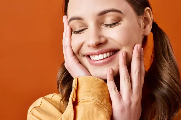 A stylish young woman in her 20s smiles brightly and covers her face with her hands in a joyful expression against an orange backdrop. — Stock Photo