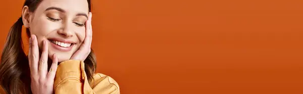 A young, stylish woman in her 20s with hands on face, deep in thought, against an orange studio background. — Stock Photo