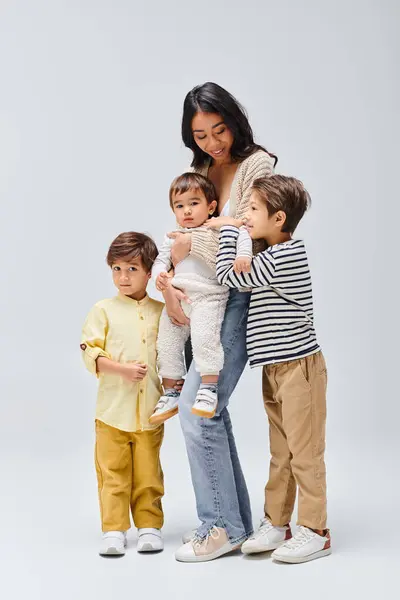 A young Asian mother tenderly holding her children, stand close by in a studio setting against a grey background. — Stock Photo