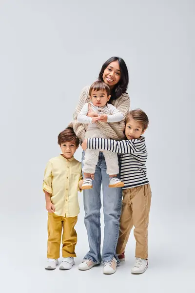 A young Asian mother stands on a grey background, holding her small children in her arms. — Stock Photo
