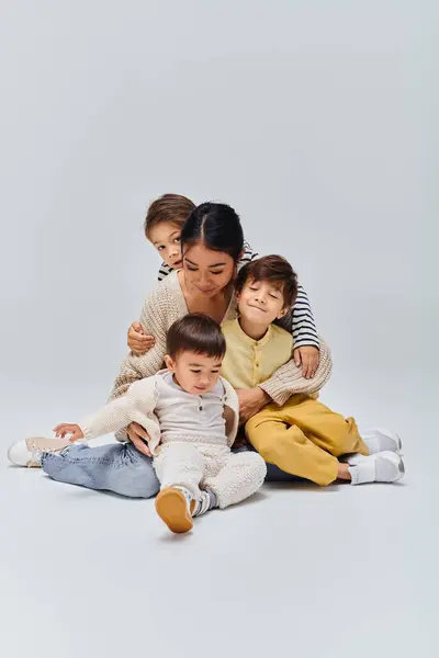 Uma jovem mãe asiática se senta no chão com seus filhos em um estúdio em um fundo cinza. — Fotografia de Stock