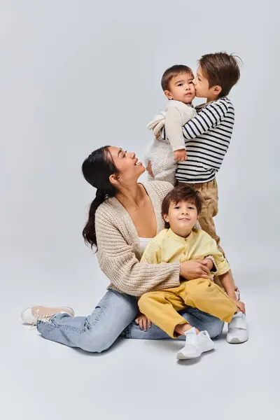 A young Asian mother sits on the ground with her children in a studio setting against a grey background. — Stock Photo