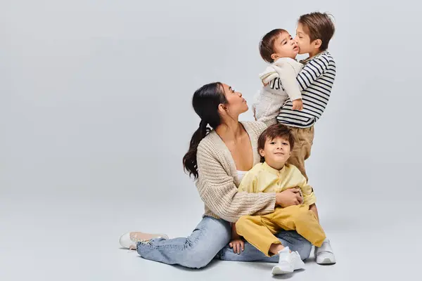 A young Asian mother sitting on the ground with her children in a studio setting against a grey background. — Stock Photo