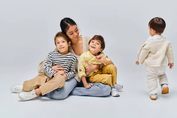 A young Asian mother sits on the ground with her children in a studio setting against a grey background, bonding closely. — Stock Photo