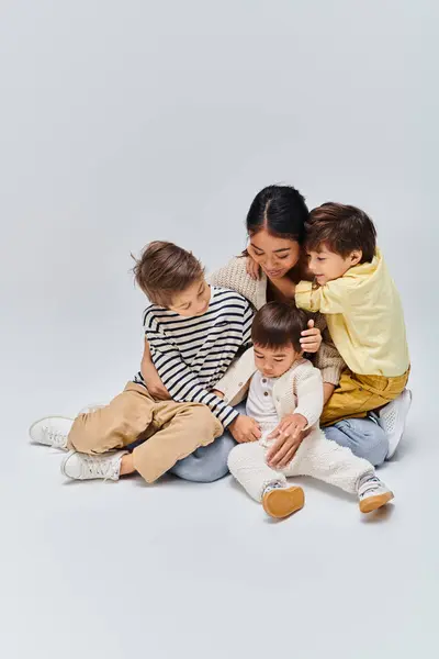 A young Asian mother sits on the ground with her children in a serene moment of bonding on a grey studio background. — Stock Photo