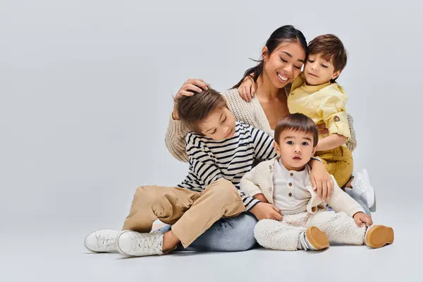 A young Asian mother sits on the floor surrounded by her children in a studio setting against a grey background. — Stock Photo