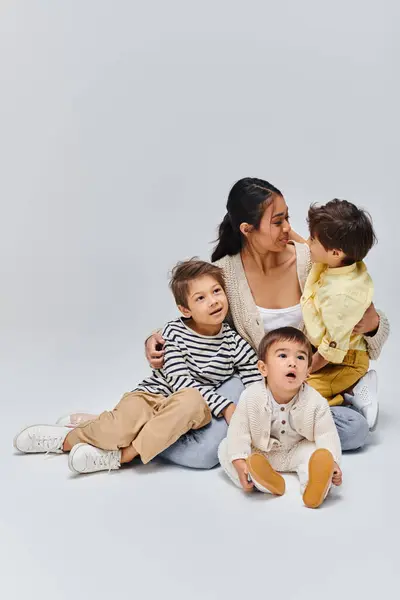 A young Asian mother sitting on the ground with her children in a serene studio setting on a grey background. — Stock Photo