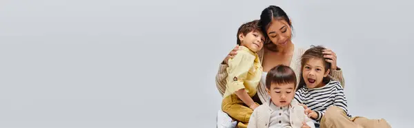 A young Asian mother sitting on a chair, lovingly embracing her children in a studio setting against a grey background. — Stock Photo