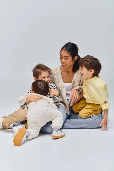 Asian mother sits on floor embracing children in studio against grey backdrop. — Stock Photo