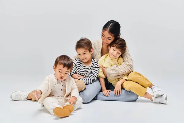 A young Asian mother sitting on the ground with her children in a studio setting on a grey background. — Stock Photo