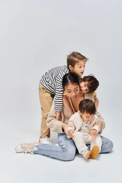 Una giovane madre asiatica e i suoi figli seduti uno sopra l'altro in uno studio sullo sfondo grigio. — Foto stock