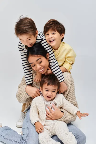 A young Asian mother and her kids create a unique human pyramid, sitting on top of each other in a studio against a grey background. — Stock Photo