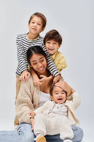 Una joven madre asiática se sienta en el suelo con sus hijos, compartiendo un cariñoso abrazo en un estudio sobre un fondo gris. - foto de stock
