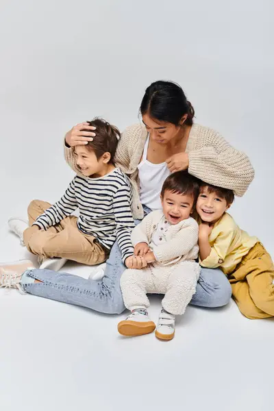 A young Asian mother is sitting on the ground with her children in a studio setting on a grey background. — Stock Photo